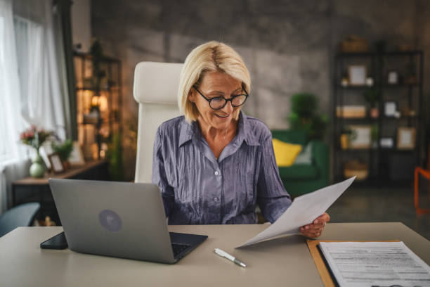 istock Mature senior woman work from home use laptop and hold documents 2156253112