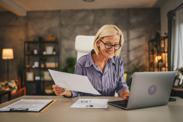 istock Mature senior woman work from home use laptop and hold documents 2156253228