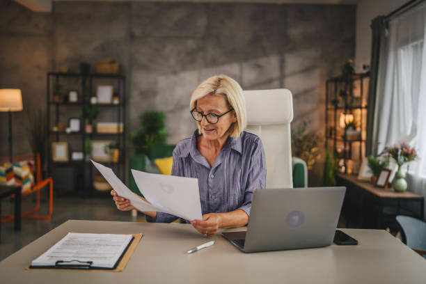 istock Mature senior woman work from home use laptop and hold documents 2156253471