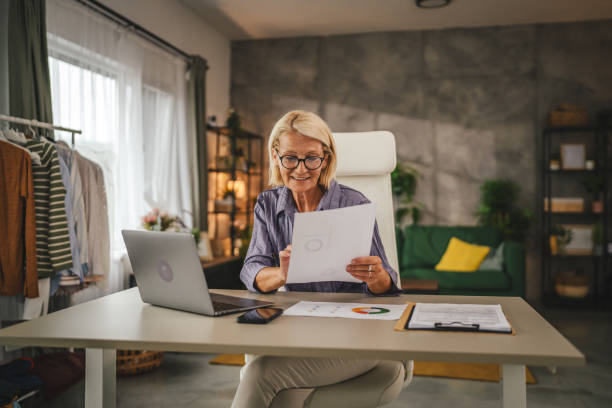 istock Mature senior woman work from home use laptop and hold documents 2156254022