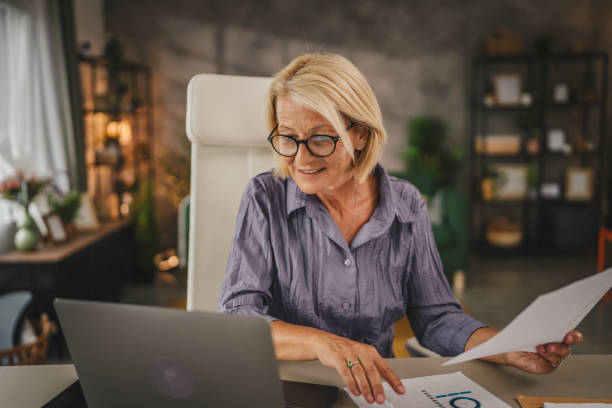 istock Mature senior woman work from home use laptop and hold documents 2156254101