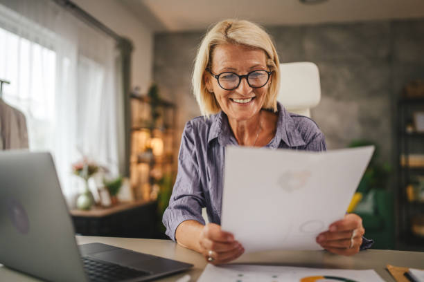 istock Mature senior woman work from home use laptop and hold documents 2156254203