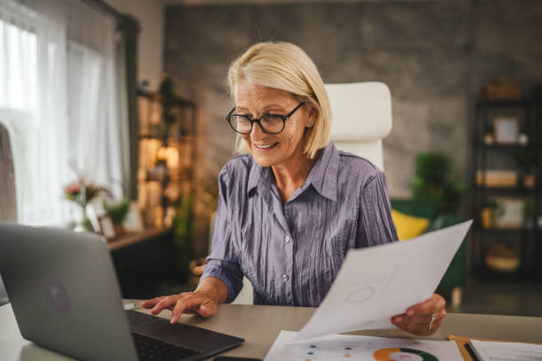 istock Mature senior woman work from home use laptop and hold documents 2156254869