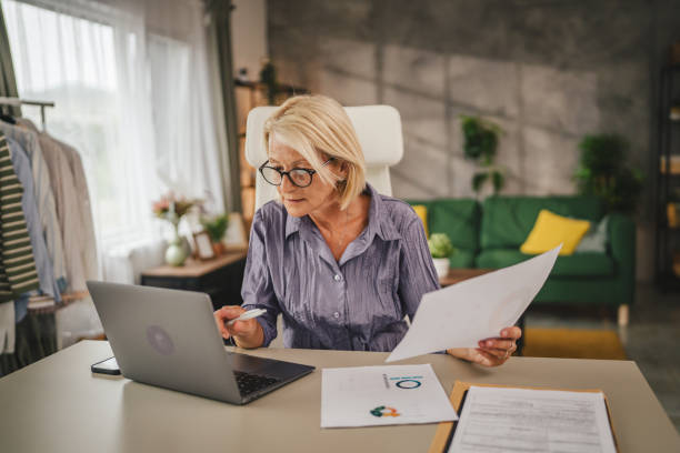 istock Mature senior woman work from home use laptop and hold documents 2156254904