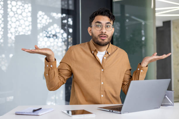 istock Confused young man in office with laptop, unsure expression and hand gesture 2156263707