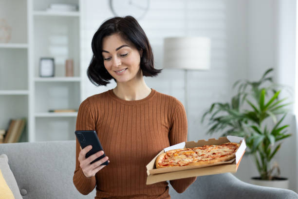 istock Woman enjoying pizza at home while browsing on smartphone 2156264722
