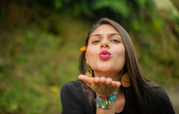 young hispanic latina woman with long hair and wearing accessories in her hands and outdoors looking straight ahead while blowing a kiss and with her hand pointing straight ahead.