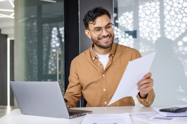 istock Smiling businessman reviewing documents at desk with laptop in modern office 2156265106