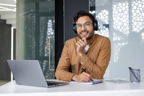 istock Smiling man with headphones working at desk with laptop in modern office setting 2156266987