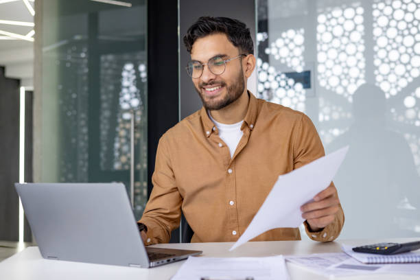 istock A smiling young Indian man is working in the office with a laptop and documents, holding papers and talking on a video call with clients and partners 2156269734