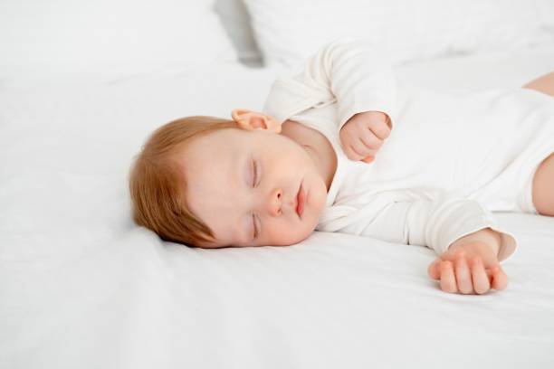 istock Baby boy sleeping under a blanket in a white bodysuit on the bed with his hand folded under his cheek, sweet healthy baby sleep at home 2156270076