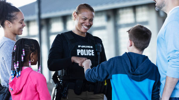 Policewoman talking to group, fist bump with boy A multiracial police woman talking to a group of parents and children outdoors. standing outside a building. She is smiling at an 11 year old boy and they are bumping fists. The view is from over the boy's shoulder. police force smiling officer stock pictures, royalty-free photos & images