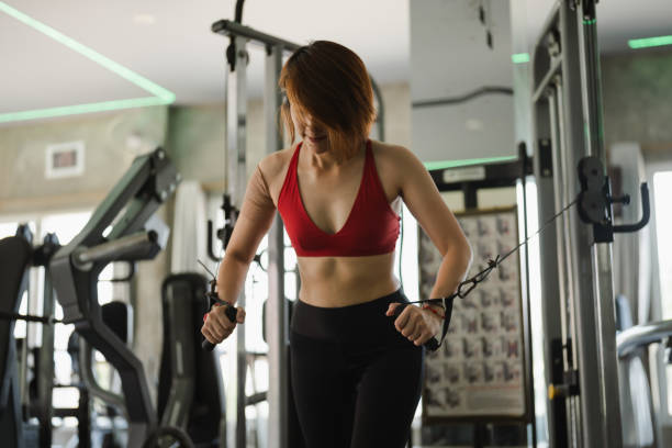 istock Asian woman is working out in a gym, lifting weights and using a cable machine. She is wearing a red top and black pants. The gym is well-equipped with various machines and equipment, including a bench 2156277232