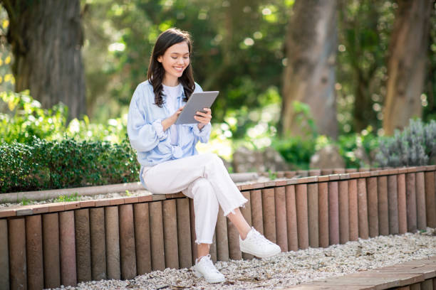 istock Engrossed young female student sitting cross-legged on a park bench, deeply focused on her tablet 2156284335