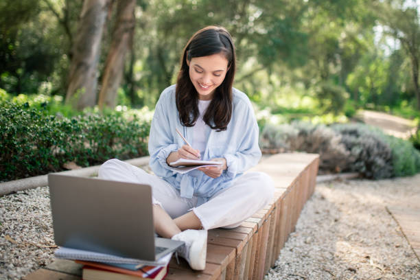A focused young woman is writing in a notebook while sitting in a tranquil park