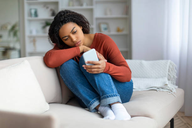 istock Young Upset Black Woman Looking At Smartphone Screen While Sitting On Couch 2156287532