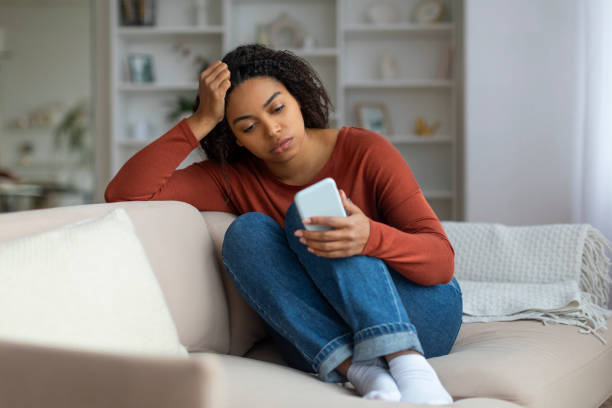 istock Bored Young Black Woman Sitting On Couch And Looking At Smartphone Screen 2156287690