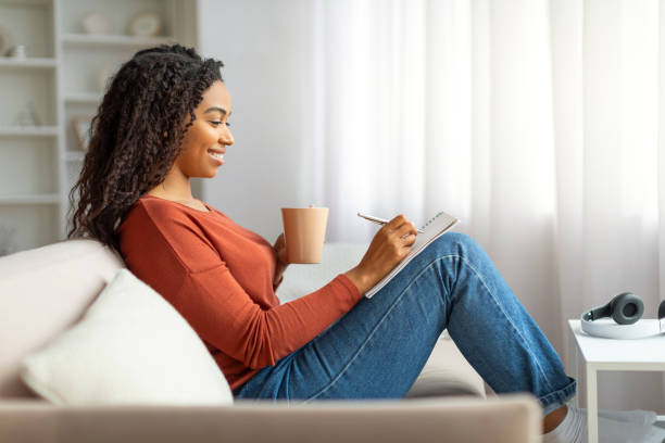 istock Black lady drinking coffee and writing in notepad while relaxing at home 2156287692