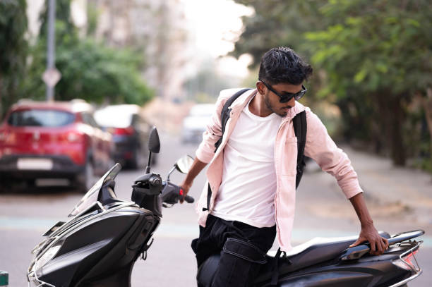 A young man rides a scooter down a city street, smiling and wearing sunglasses. He is dressed casually in a light pink shirt and white t-shirt, with a backpack on his shoulders