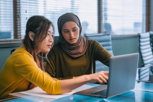 Two Asian businesswomen talking while working together with laptop in a modern office startup