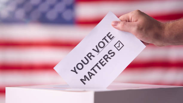 Hand Placing Ballot in Box With American Flag in Background