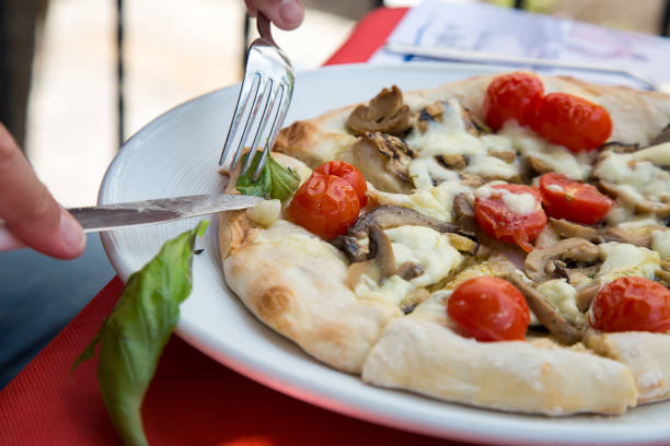 A person is using a fork to slice a pizza. The scene involves food, tableware, ingredients, a recipe, a plate, fast food, kitchen utensils, staple food, produce, and a dish