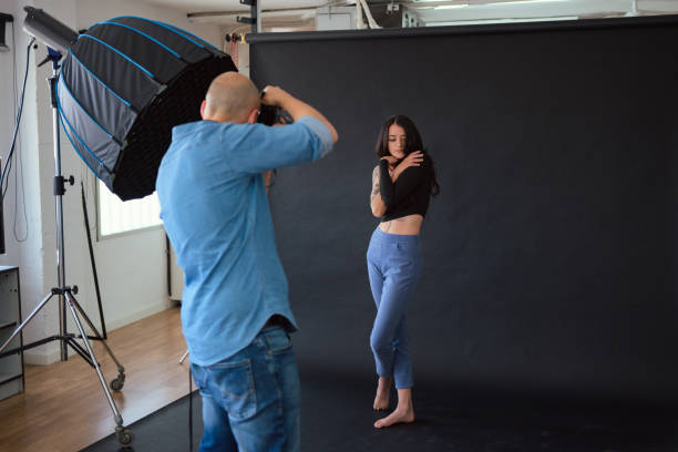 A photographer shoots a female model in a studio. The model is standing, wearing a black top and blue jeans. The photographer is holding a camera. The background is a black backdrop a with softbox