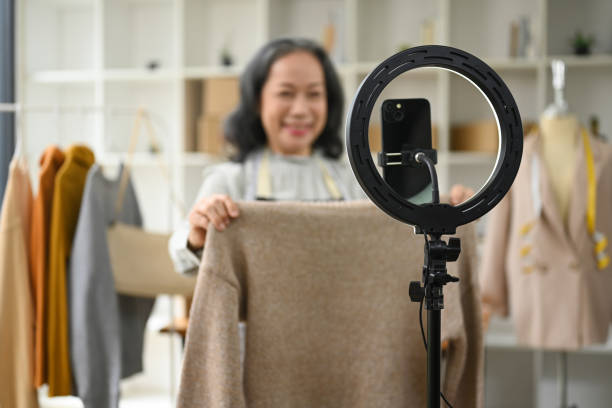 Smiling middle age woman showing clothes in front of smartphone camera while live streaming at her shop