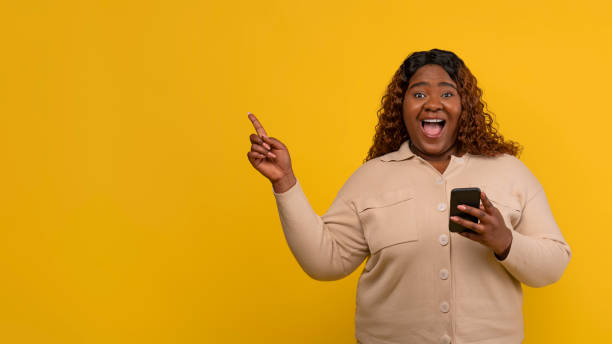 istock Excited chubby african american lady with smartphone pointing copy space 2156324213