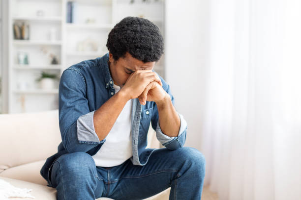 istock Distressed Young Black Man Seated on Couch With Hands Covering Face 2156332349