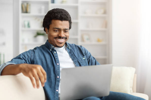 istock Smiling Black Man Using Laptop on Couch in Bright Living Room 2156332378