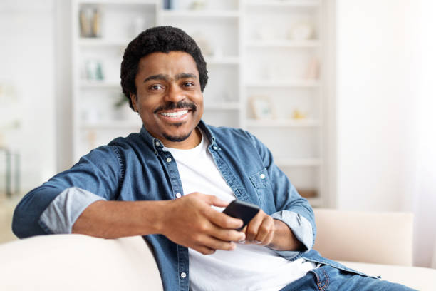 istock Smiling Black Man Relaxing on Couch With Smartphone in Bright Living Room 2156332690