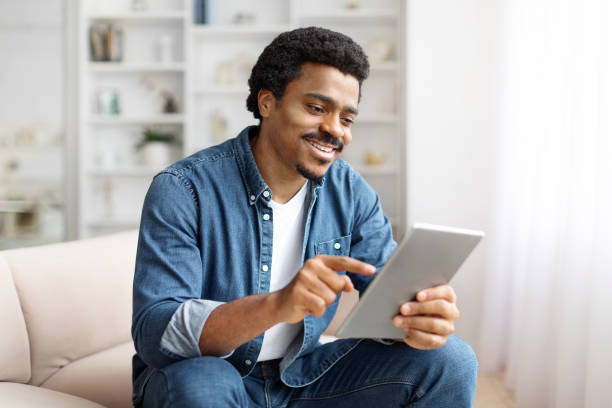 istock Smiling Black Man Using Digital Tablet On Couch in Bright Living Room 2156332697