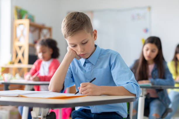 istock Sad schoolboy sitting at desk, looking down, leaning head on hand and writing in copybook during lesson or test 2156338450