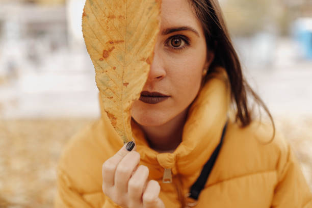 Charming young lady in warm jacket holding huge yellow leaf near eye and looking at camera while standing on blurred background of city street on autumn day