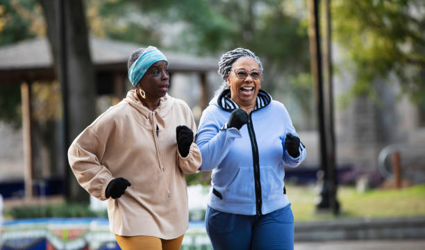 Two black senior women exercising, power walking in park Two senior women exercising together, jogging or power walking in a city park. The African-American woman in the light blue sweatshirt is in her 70s. Her friend running beside her is in her 60s. short weave hairstyles stock pictures, royalty-free photos & images
