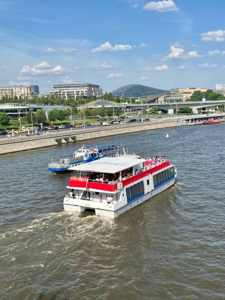 Moscow, Russia - May 17, 2025: A river tram sails from Filevsky Park along  Moscow River past the Red Bridge on  cloudy spring day, demonstrating  unity of nature, transport, architecture, and urban space.