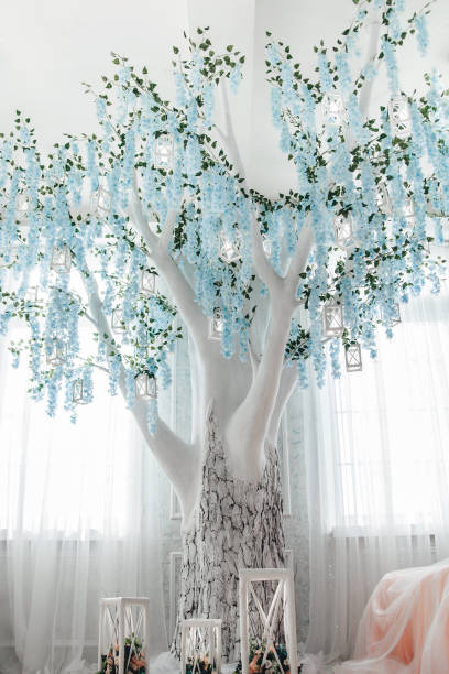 Decorative white tree with cascading blue flowers and hanging lanterns in white room. Soft natural light enhances serene and magical ambiance. Sheer curtains and delicate furnishings complement scene