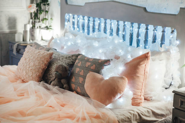 Elegant bedroom setup featuring pastel bedding, heart-shaped pillows, and a teddy bear. Soft string lights adorn headboard, creating warm ambiance. Nightstand with plant and decor adds to coziness