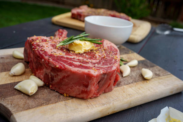 Raw Tomahawk Rib Eye Beef Steak on a Rustic Wooden Cutting Board Ready to Cook Next to a Charcoal Grill in a Residential Back Yard in Summertime