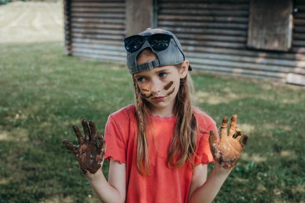 A young girl in a backward cap shows her muddy face and hands to the camera while playing in the park.