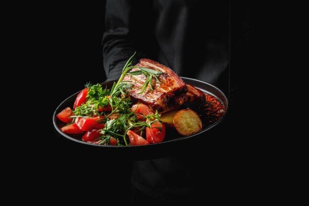 istock A plate of seared steak garnished with herbs, accompanied by sliced tomatoes and greens, held by a person against a dark background 2156397439