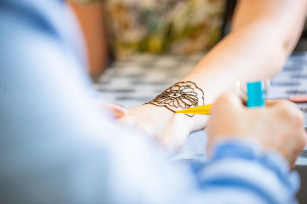 A henna artist carefully applies an intricate design to a womans wrist using a small cone filled with henna paste. The womans hand is resting on a table, and the artists hand is visible in the foreground. The image is captured from a close-up perspective, focusing on the detailed henna application process.