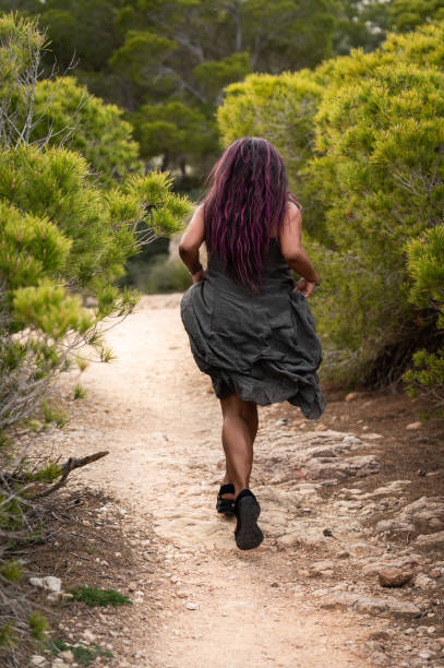 Vertical rear view of a latin beauty woman walking along a forest footpath playing with long dress