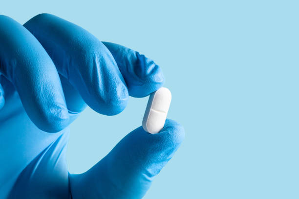istock Blue-gloved hand holding a white pill against a light blue background. 2156456915