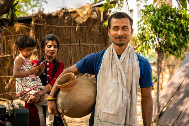 Portrait of happy traditional, rural indian young man carrying drinking water clay pot and contemplating while looking away and her wife stands behind with child in the village.