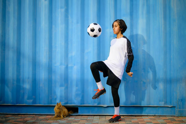 Full length of skilled teenage female player playing with soccer ball. Female athlete is practicing against blue wall. She is wearing sports uniform.