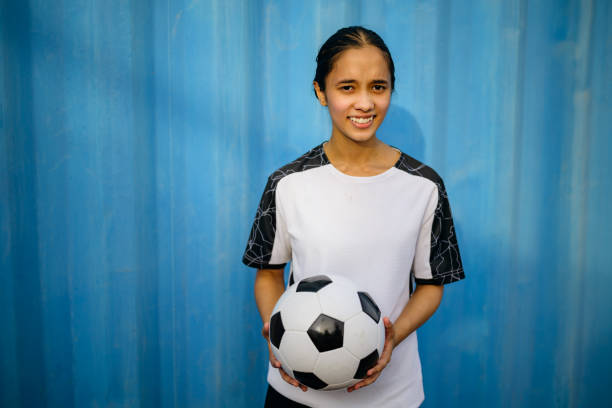 Portrait of smiling teenage soccer player with ball. Female athlete is standing against blue wall. She is wearing sports jersey.