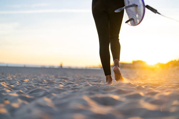 A cheerful Black woman in a sleek wetsuit strides across a sandy beach, surfboard in hand, with the glow of the setting sun highlighting her silhouette.