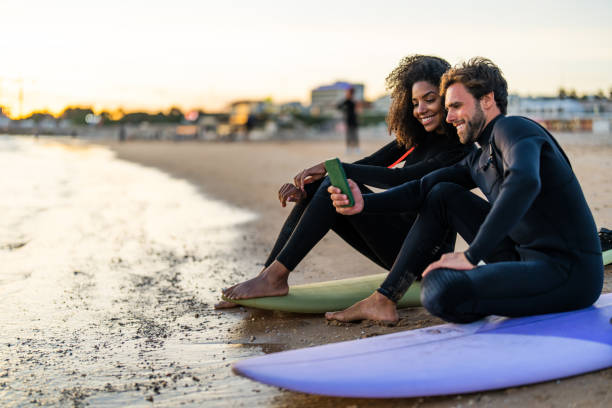 A smiling Black woman and Caucasian man in wetsuits sit on colorful surfboards, using a smartphone for a selfie. Golden hour light adds warmth to the sandy shoreline setting.